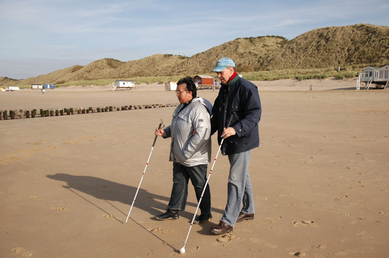 Twee mensen die blind of slechtziend zijn lopen samen op het strand. Ze worden door de Robert Coppes Stichting ondersteund om het leven te leiden dat bij hen past.