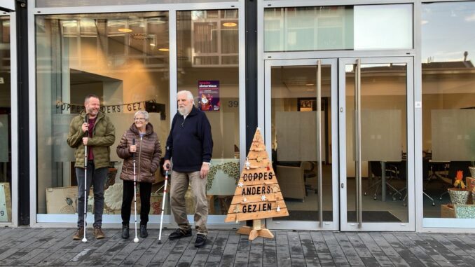 Drie cliënten staan voor dagbesteding Coppes Anders Gezien in Geleen. Ze lachen en dragen een blindenstok. 