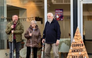 Drie clienten staan voor de nieuwe locatie van dagbesteding Coppes Anders Gezien. Ze lachen en dragen een blindenstok.