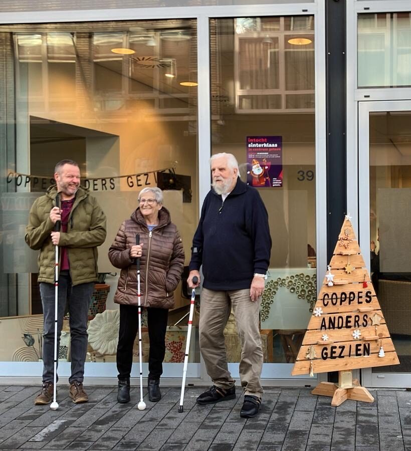 Drie clienten staan voor de nieuwe locatie van dagbesteding Coppes Anders Gezien. Ze lachen en dragen een blindenstok.