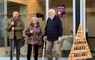 Drie clienten staan voor de nieuwe locatie van dagbesteding Coppes Anders Gezien. Ze lachen en dragen een blindenstok.