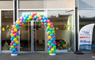 Foto van de voordeur van dagbestedingslocatie Coppes Anders Gezien in Geleen. Voor de glazen deur staat een ballonnenboog met ballonnen in alle kleuren van de regenboog. Naast de deur staat een vlag met het logo van de Robert Coppes Stichting en 'Welkom'.