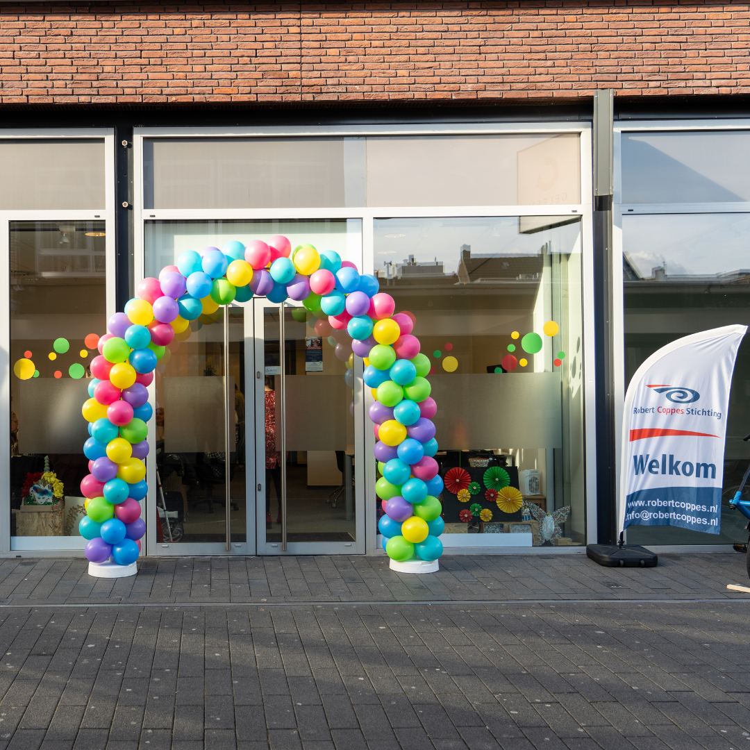 Foto van de voordeur van dagbestedingslocatie Coppes Anders Gezien in Geleen. Voor de glazen deur staat een ballonnenboog met ballonnen in alle kleuren van de regenboog. Naast de deur staat een vlag met het logo van de Robert Coppes Stichting en 'Welkom'.