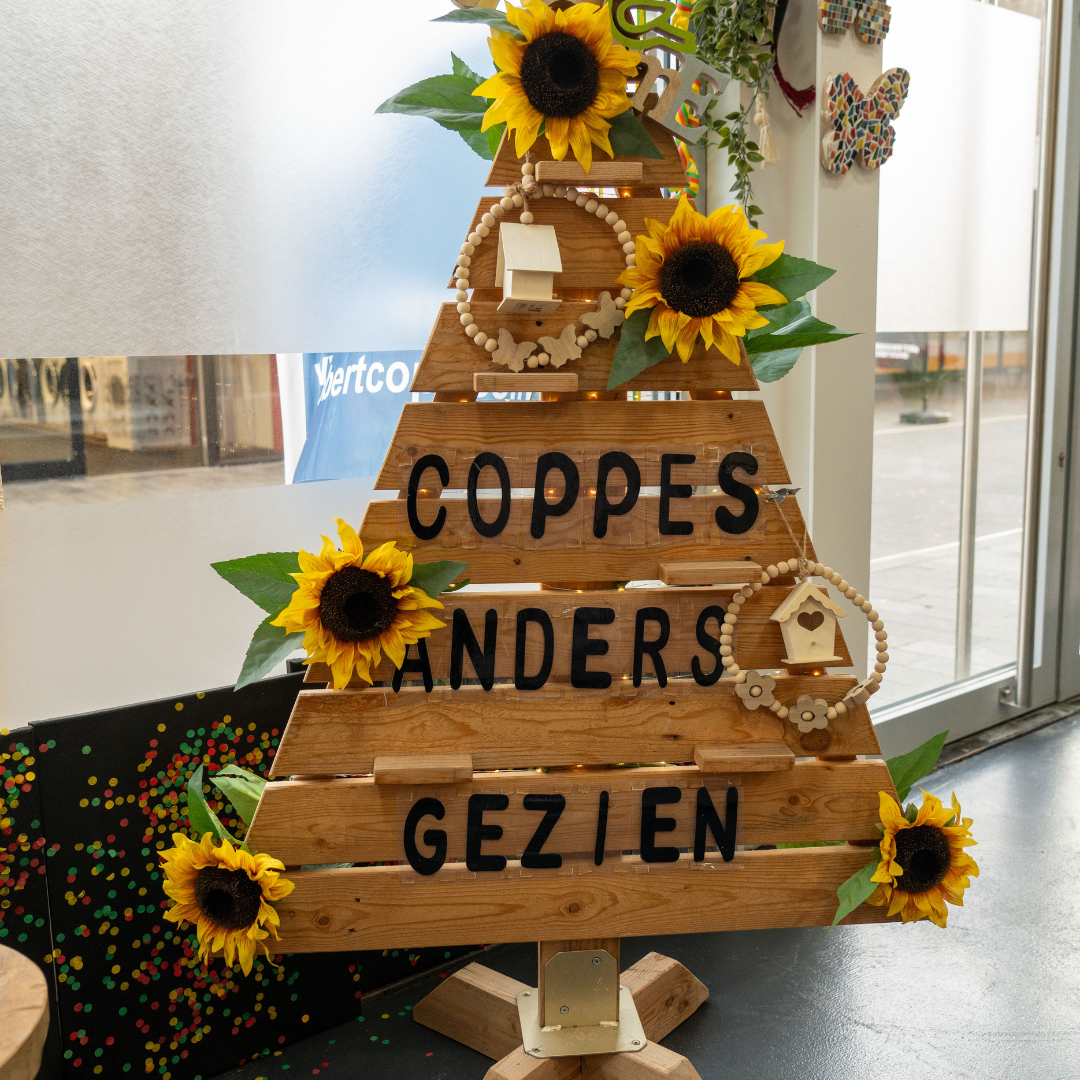 Een foto met een houten bord in de vorm van een driehoek met daarop Coppes Anders Gezien. Het bord is versierd met zonnebloemen.