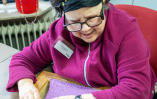 Een vrouw met een roze vest, een zwarte haarband en een bril weeft een handwerk met lila garen. Ze zit op een rode stoel aan een grijze tafel. Op de achtergrond staat een rode pot op een vensterbank boven een radiator.