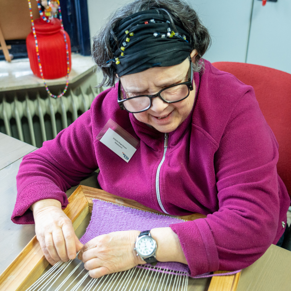 Een vrouw met een roze vest, een zwarte haarband en een bril weeft een handwerk met lila garen. Ze zit op een rode stoel aan een grijze tafel. Op de achtergrond staat een rode pot op een vensterbank boven een radiator.
