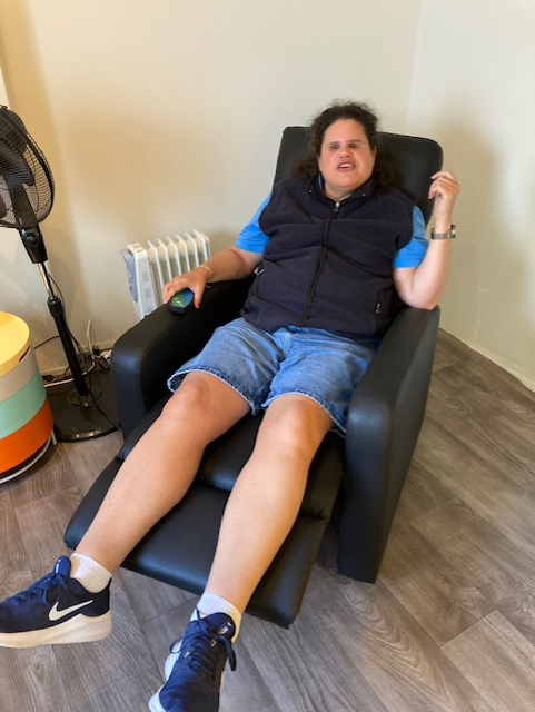 Een vrouw met een zwarte bodywarmer, blauw T-shirt en een korte spijkerbroek zit in een zwarte massagestoel. Naast de stoel staat een zwarte ventilator en een gekleurd kastje.