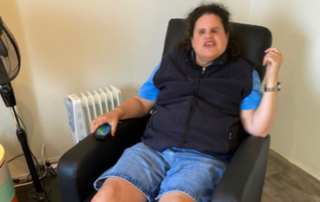 Een vrouw met een zwarte bodywarmer, blauw T-shirt en een korte spijkerbroek zit in een zwarte massagestoel. Naast de stoel staat een zwarte ventilator en een gekleurd kastje.