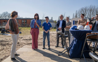 Bestuurder Marian van Heerebeek (RCS) spreekt de aanwezigen toe tijdens de feestelijke start van de bouw aan de Hertoglaan. Naast haar staat cliënt en lid van de centrale cliëntenraad Marius, en op de achtergrond luisteren genodigden mee.
