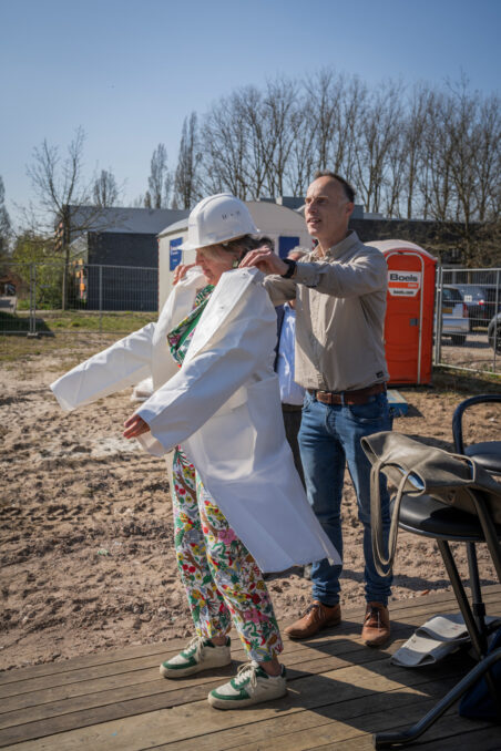 Start bouw Hertoglaan Wethouder Yvonne Vos krijgt hulp bij het aantrekken van een bouwhelm en jas, klaar om het startsein te geven voor de bouwwerkzaamheden.