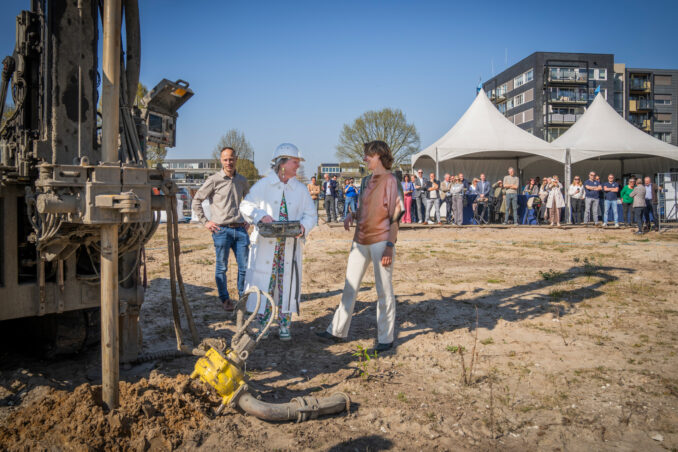 Start bouw Hertoglaan Wethouder Yvonne Vos bedient de machine voor de bronboring, waarmee zij officieel het startsein geeft voor de bouw. Omstanders kijken toe en applaudisseren.
