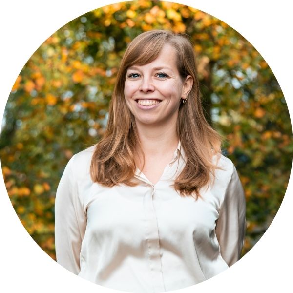 Profielfoto van Edine van Munster, onderzoeker bij het project eenzaamheid in Zicht. Edine heeft lang goudblond haar dat over haar schouders valt, draagt een glanzende witte blouse en kijkt lachend naar de camera. Ze is buiten gefotografeerd voor bomen met herfstkleuren.