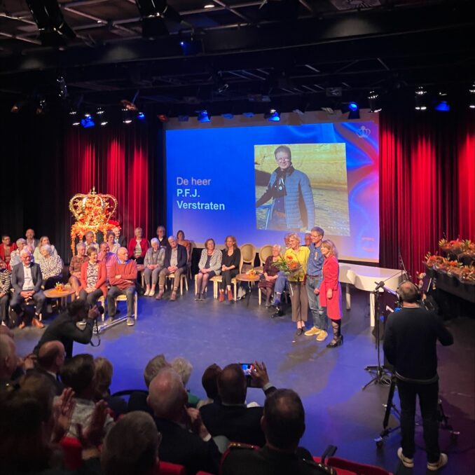 Peter_linjte_burgermeester Peter staat met zijn vrouw en de burgemeester op het podium tijdens de officiële lintjesuitreiking. Op de achtergrond zit een groep genodigden en is op een groot scherm zijn naam te lezen: “De heer P.F.J. Verstraten”, met een foto van hemzelf ernaast.