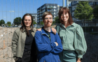 Foto uit het Brabants Dagblad. Drie personen staan naast elkaar voor een bouwterrein, met een hek op de achtergrond en moderne gebouwen erachter. Eén persoon draagt een blauwe jas en staat met de armen over elkaar, de twee andere personen staan ernaast. Op de achtergrond is een open, zanderig terrein te zien onder een heldere blauwe lucht.