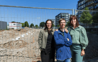 Drie personen stan voor het hek van een bouwplaats waar nieuwe woningen voor de Robert Coppes Stichting worden gebouwd. Het zijn Mariëlle Huffmeijer, financieel adviseur vastgoed, een bewoner en bestuurder Marian van Heerebeek.