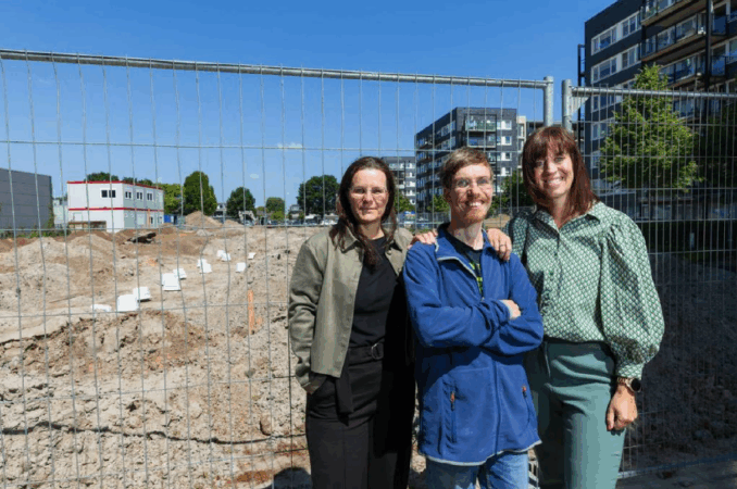 foto bd Drie personen stan voor het hek van een bouwplaats waar nieuwe woningen voor de Robert Coppes Stichting worden gebouwd. Het zijn Mariëlle Huffmeijer, financieel adviseur vastgoed, een bewoner en bestuurder Marian van Heerebeek.