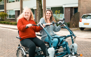 Twee vrouwen fietsen samen op een duofiets in een woonwijk. Ze lachen en kijken voor zich uit. De vrouw links draagt een rode trui, de vrouw rechts een grijze. Op de achtergrond zijn huizen, een boom en een geparkeerde auto zichtbaar.