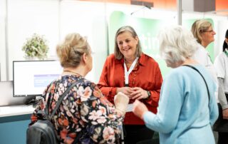 Een vrouw met een rode blouse staat lachend in gesprek met twee oudere vrouwen op een beurs of informatiemarkt. Op de achtergrond zijn een computerscherm, een groene banner en andere aanwezigen zichtbaar.