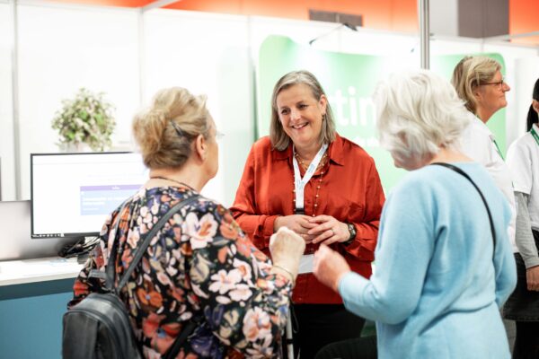 Een vrouw met een rode blouse staat lachend in gesprek met twee oudere vrouwen op een beurs of informatiemarkt. Op de achtergrond zijn een computerscherm, een groene banner en andere aanwezigen zichtbaar.