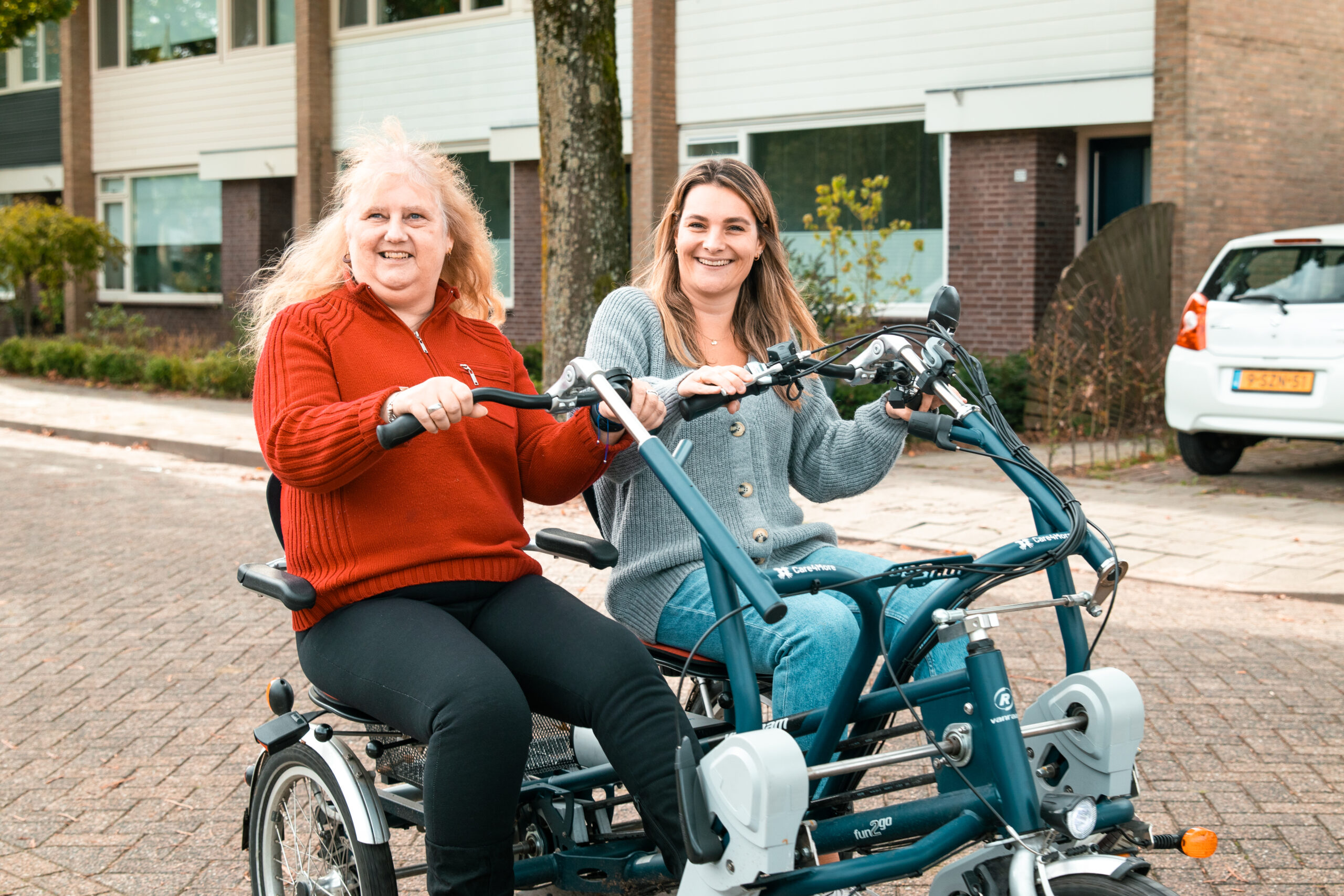Twee vrouwen zitten samen op een duofiets in een woonwijk. Ze lachen en genieten van het fietsen; de vrouw links draagt een rode trui en de vrouw rechts een grijze vest.