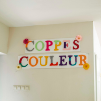 Twee witte borden aan een muur met daarop de woorden “COPPES COULEUR” in grote, kleurrijke letters van wol. Rond de letters zijn enkele kunstbloemen bevestigd. Onder de borden zijn lichtschakelaars zichtbaar en rechts een deuropening.