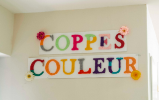 Twee witte borden aan een muur met daarop de woorden “COPPES COULEUR” in grote, kleurrijke letters van wol. Rond de letters zijn enkele kunstbloemen bevestigd. Onder de borden zijn lichtschakelaars zichtbaar en rechts een deuropening.