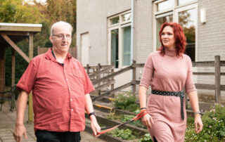 Een cliënt en een begeleider lopen samen buiten langs een gebouw. De cliënt draagt een rode blouse en houdt een rood leidsel vast, de begeleider in een roze jurk loopt naast hem en ondersteunt hem tijdens het lopen.