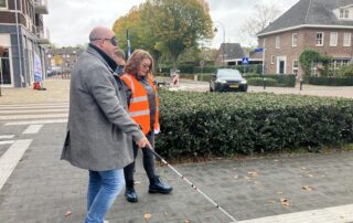 Persoon met een grijze jas en oogmasker loopt met een witte taststok over een trottoir, begeleid door iemand in een feloranje veiligheidsvest. Op de achtergrond zijn struiken, een geparkeerde auto en huizen zichtbaar.