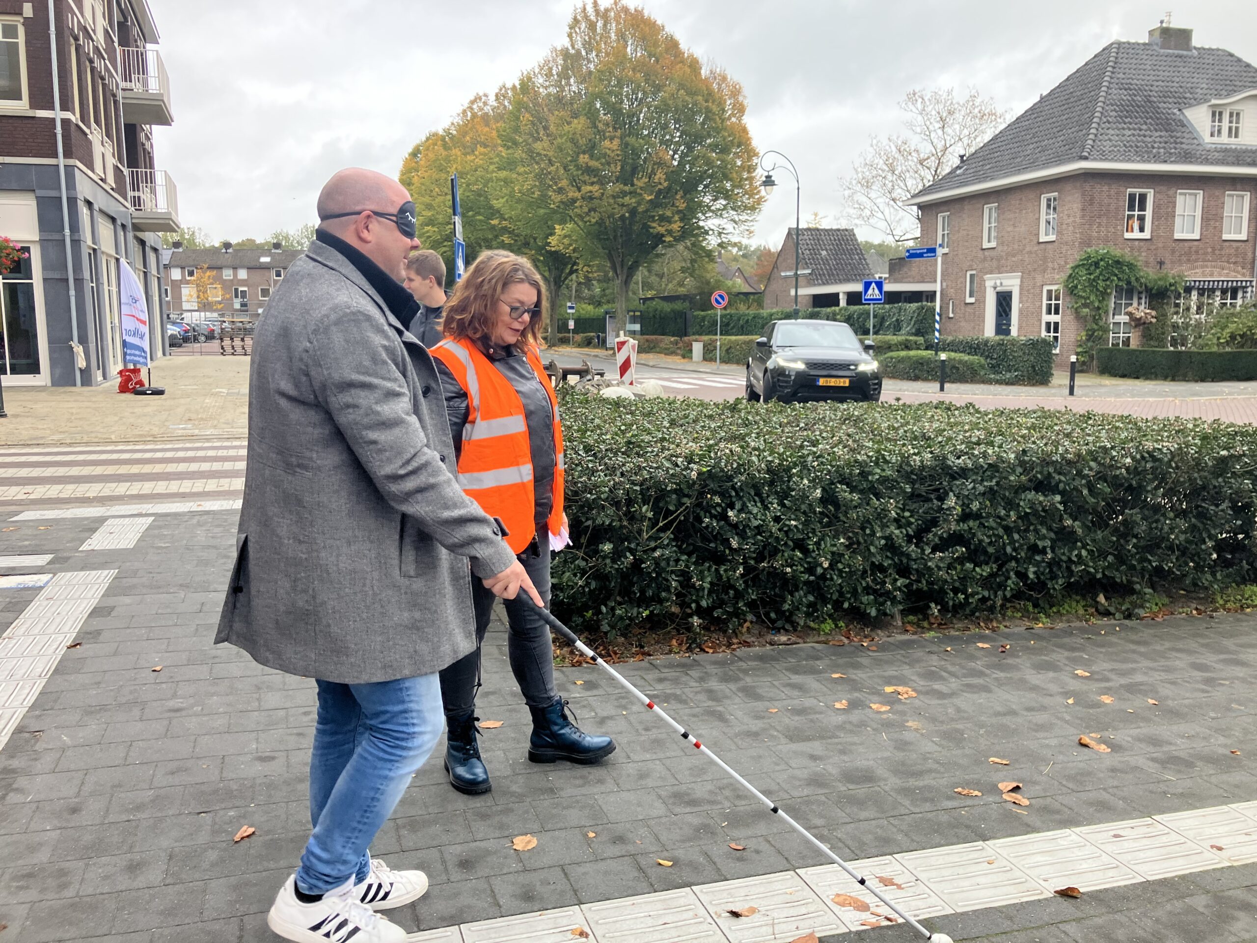 Persoon met een grijze jas en oogmasker loopt met een witte taststok over een trottoir, begeleid door iemand in een feloranje veiligheidsvest. Op de achtergrond zijn struiken, een geparkeerde auto en huizen zichtbaar.