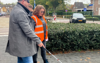 Foto van twee personen die samen over een stoep lopen in een woonwijk in Vught, tijdens de Dag van de Witte Stok 2025. Eén persoon gebruikt een witte stok en draagt een oogmasker, de andere persoon loopt ernaast en draagt een oranje veiligheidshesje. Op de achtergrond zijn geparkeerde auto’s, huizen en bomen zichtbaar.