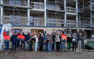 Een grote groep mensen staat voor een appartementencomplex in aanbouw. Ze lachen en steken juichend hun armen in de lucht. Tussen de groep staan bouwmedewerkers in feloranje jassen en toekomstige bewoners en betrokkenen, voor een gevel met steigers.
