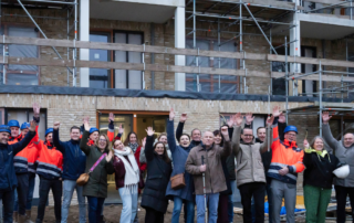 Groep mensen staat voor een appartementencomplex in aanbouw in Vught en steekt de armen omhoog. Op de achtergrond zijn steigers en balkons zichtbaar. Enkele personen dragen bouwhelmen en veiligheidshesjes. Eén persoon vooraan staat met een witte stok.