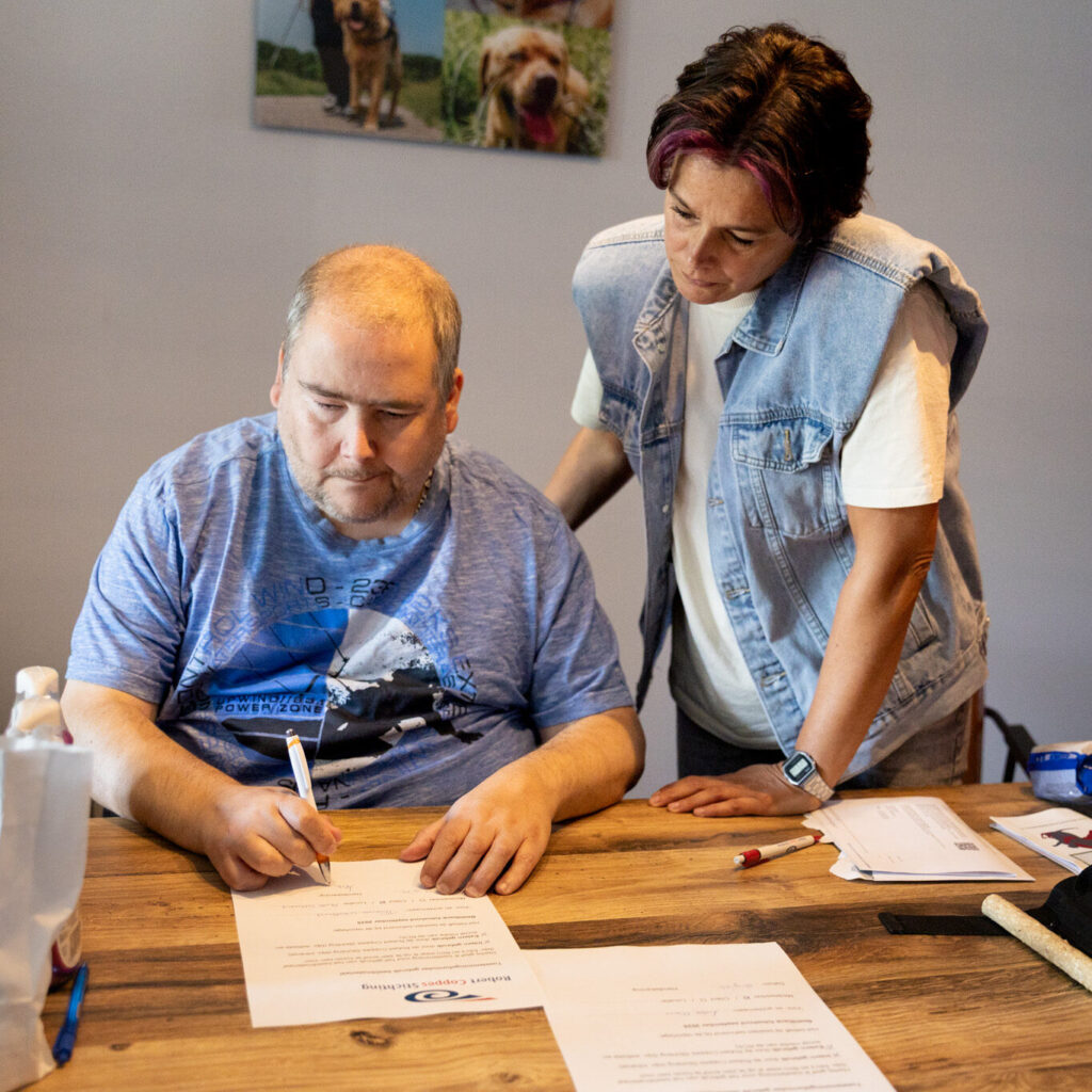 rcs mid resolutie-77 Een man schrijft aan tafel terwijl de begeleider naast hem staat en meekijkt.