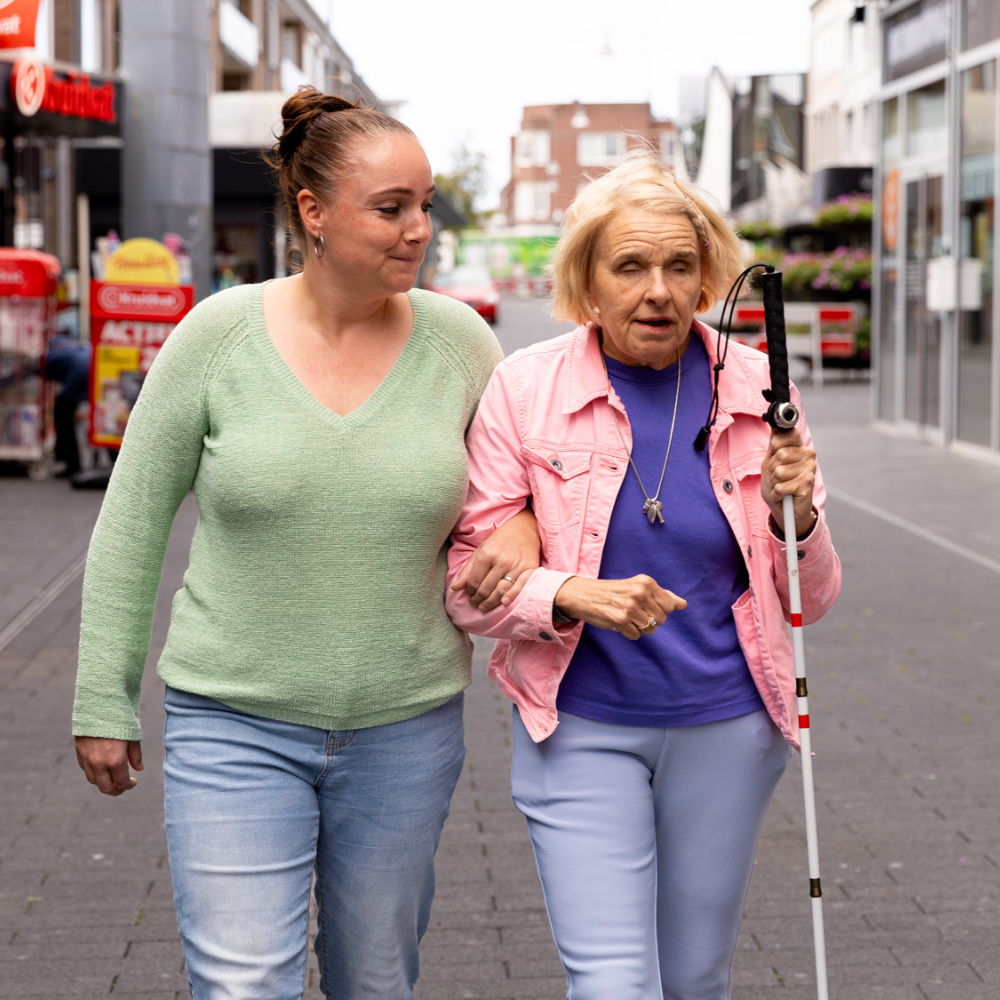 Beeld website – 1000×1000 – dagbesteding v2 Twee personen lopen arm in arm door een winkelstraat; één van hen gebruikt een witte taststok.