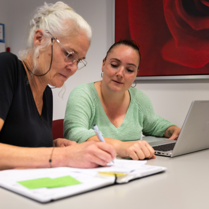 Beeld website – 1000×1000 – overleg Twee personen zitten aan tafel en werken samen; één maakt aantekeningen terwijl de ander op een laptop werkt in een rustige vergaderruimte.