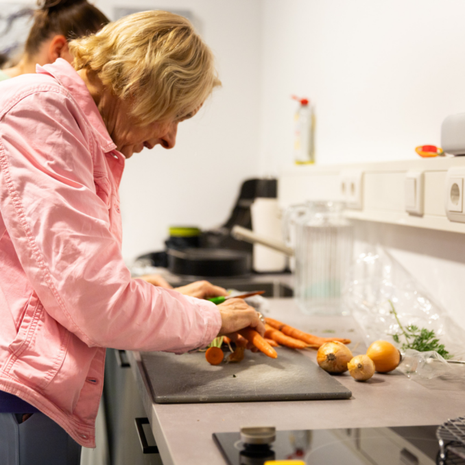 Beeld website – 1000×1000 – wortels snijden Een persoon snijdt wortels op een snijplank in een keuken, terwijl op het aanrecht ook andere groenten klaarliggen.