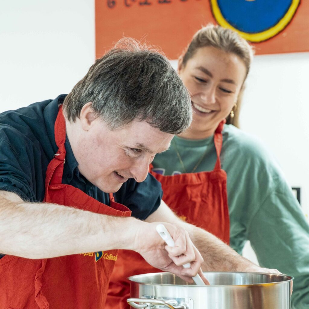 Dagbesteding 9 Een man roert in een pan terwijl de begeleider naast hem staat en glimlacht.