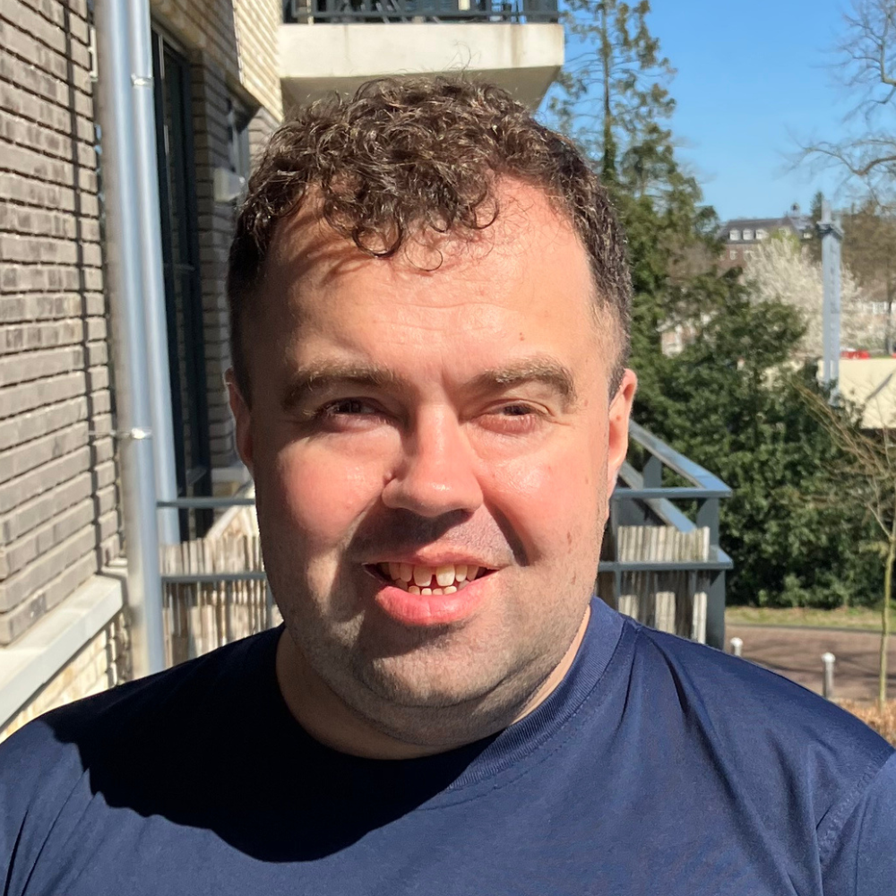Persoon met kort donker krullend haar, staand buiten voor een gebouw met balkon, met bomen op de achtergrond.