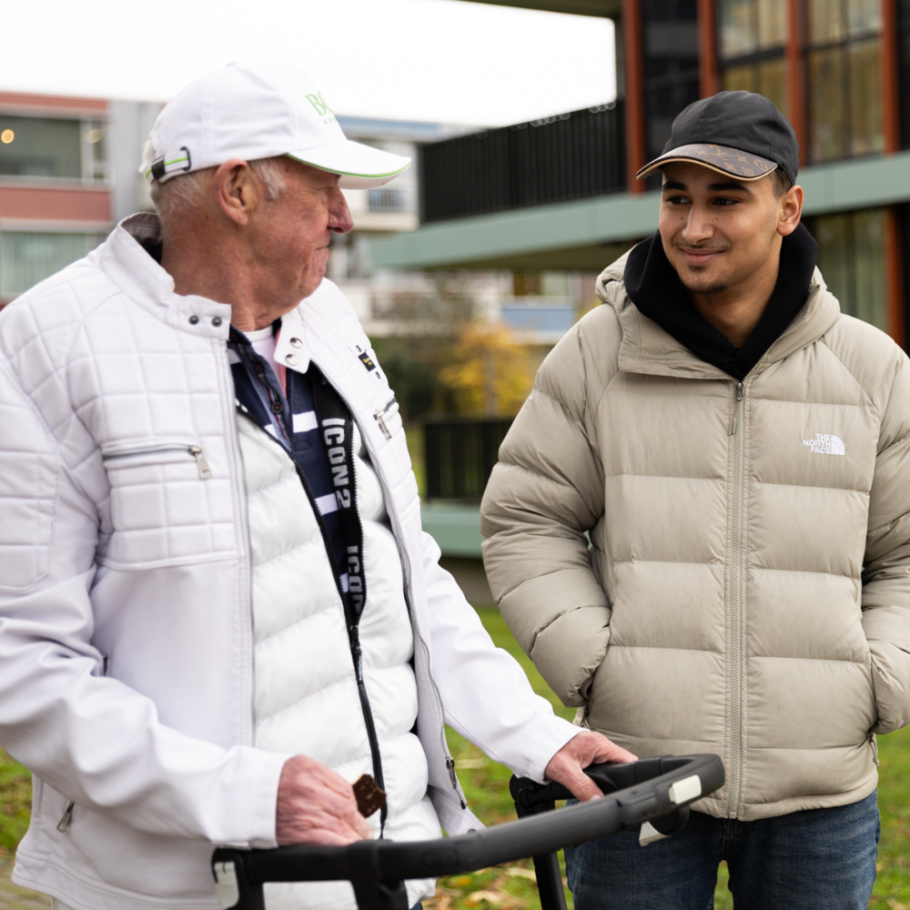 Een cliënt gebruikt een rollator terwijl een begeleider naast hem staat in een buitenomgeving.