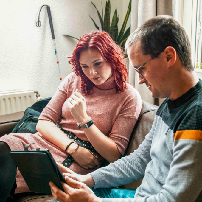 Een cliënt en een begeleider zitten samen op een bank en bekijken informatie op een tablet.