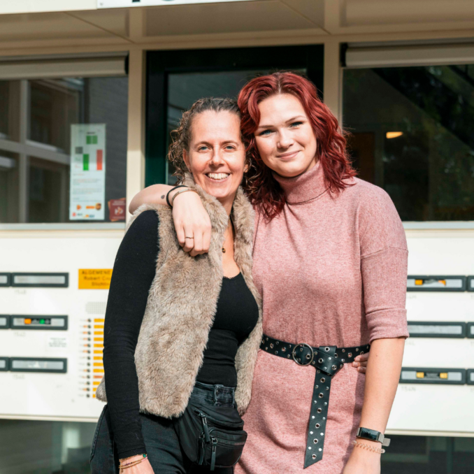 Website – collega begl – 1000×1000 Twee collega’s staan buiten voor een gebouw en hebben hun arm om elkaar heen.