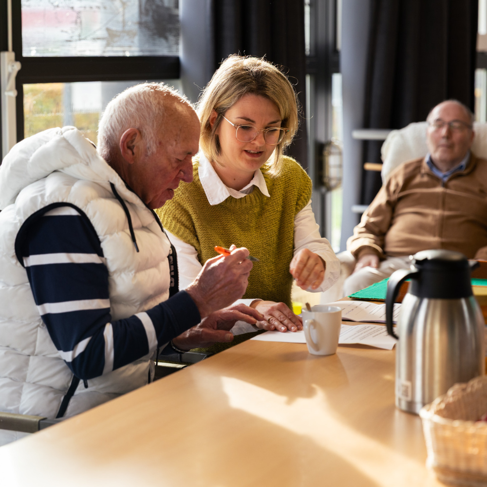 Twee personen zitten naast elkaar aan een tafel. Ze werken samen aan een document, met een beker op tafel.