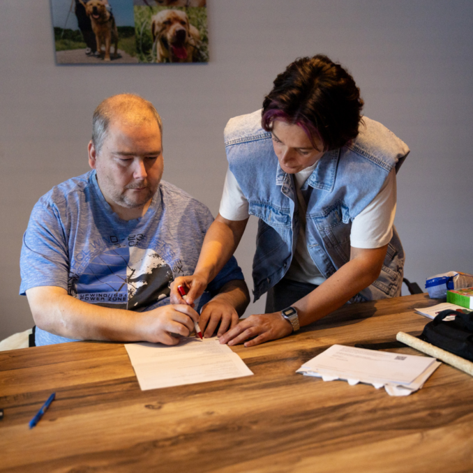 Website – papierentekenen awb – 1000×1000 Een begeleider helpt een cliënt bij het ondertekenen van een document aan een tafel.