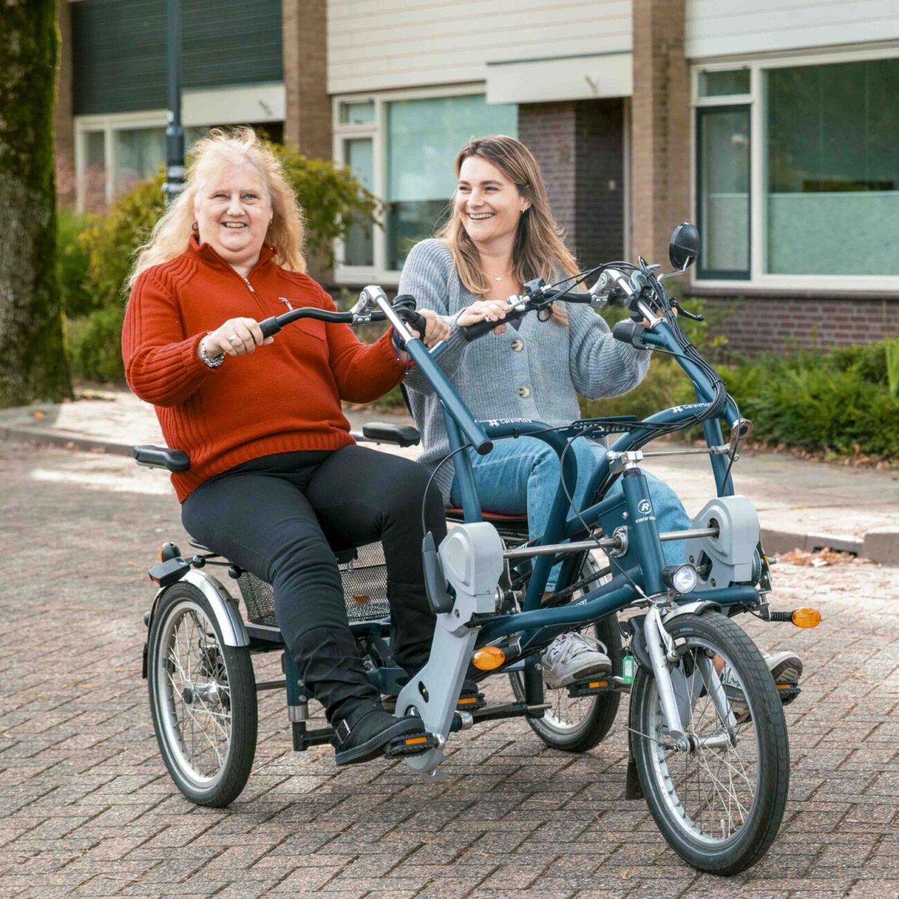 Wonen ZW duofiets 6 Twee vrouwen zitten samen op een duofiets en fietsen door de straat. De ene vrouw kijkt lachend in de camera, de andere vrouw kijkt haar lachend aan. Op de achtergrond een rij huizen.