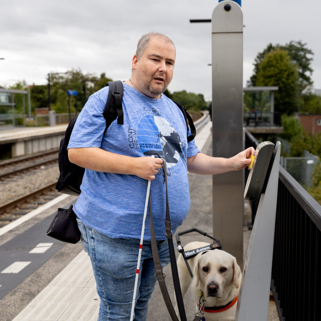 rcs mid resolutie-107 Een man staat op een perron met een geleidestok en zijn hond en checkt zijn OV-kaart in bij een incheckpaal.