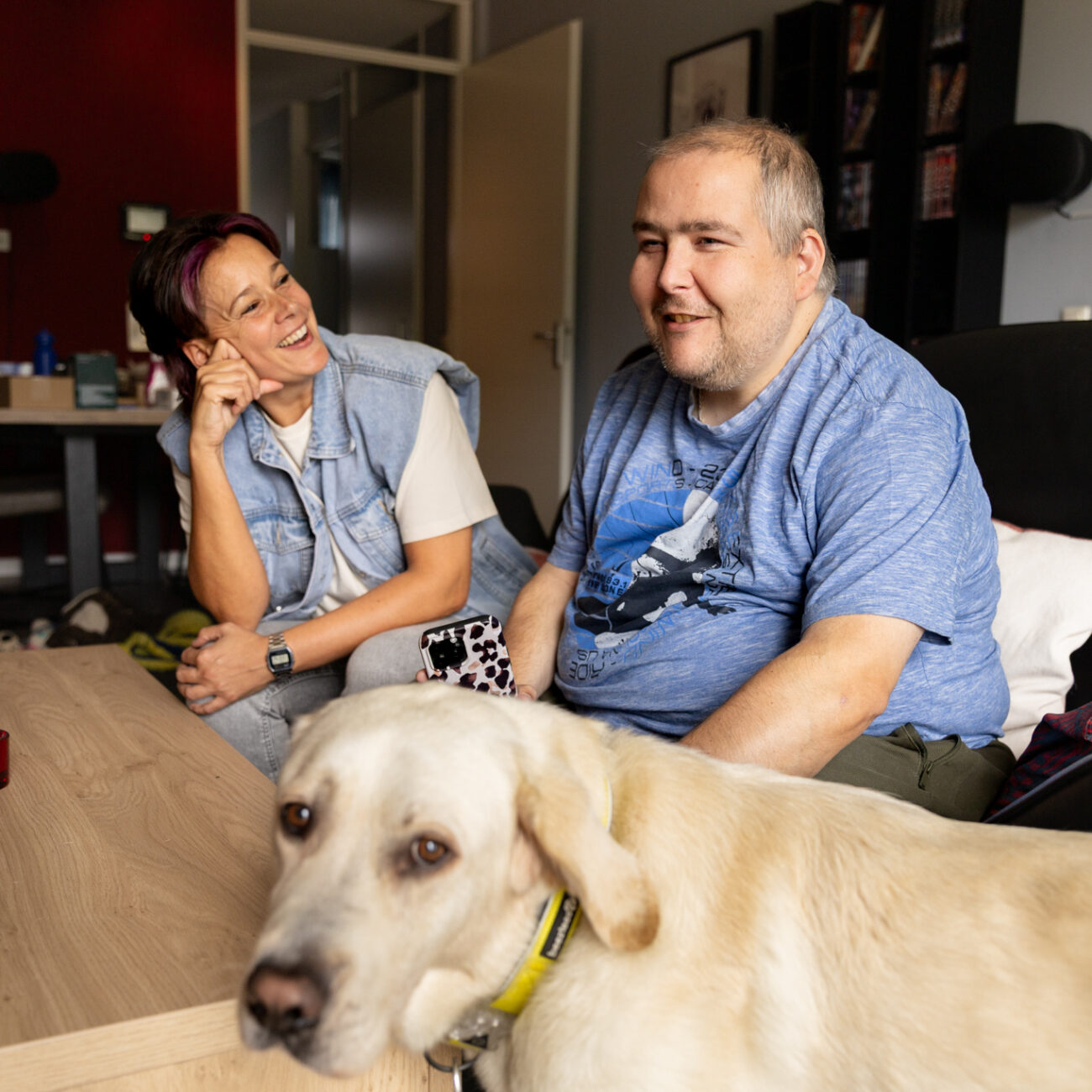 rcs mid resolutie-84 Een man zit op de bank en lacht breed. Naast hem zit zijn begeleider, zij kijkt hem lachend aan. Op de voorgrond kijkt een blindegeleidenhond naar de camera.