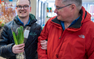 Twee personen lopen samen voor een winkel. De persoon links houdt een bos bloemen vast. De persoon rechts draagt een rode jas en houdt een product vast. De personen lopen dicht naast elkaar en raken elkaars arm aan.