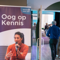 Binnenruimte met een banner van “Oog op Kennis” van Kennis over Zien. Op de banner staat een foto van een spreker met een microfoon. Naast de banner is een doorgang naar een zaal waar meerdere mensen rondlopen en met elkaar in gesprek zijn.
