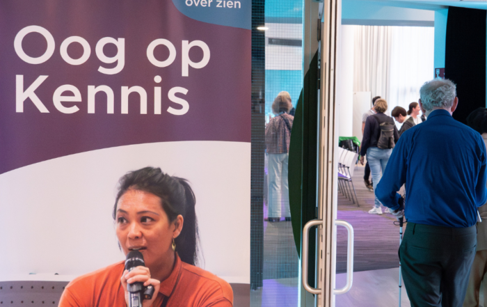 Binnenruimte met een banner van “Oog op Kennis” van Kennis over Zien. Op de banner staat een foto van een spreker met een microfoon. Naast de banner is een doorgang naar een zaal waar meerdere mensen rondlopen en met elkaar in gesprek zijn.