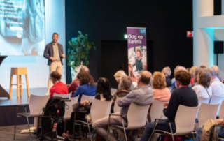 Bezoekers luisteren naar een spreker tijdens het kennisevent Oog op Kennis. De spreker staat op een podium voor een presentatie, terwijl deelnemers aandachtig in de zaal zitten. Op de achtergrond staat een banner van Oog op Kennis. Voorin ligt een hulphond rustig op de grond.